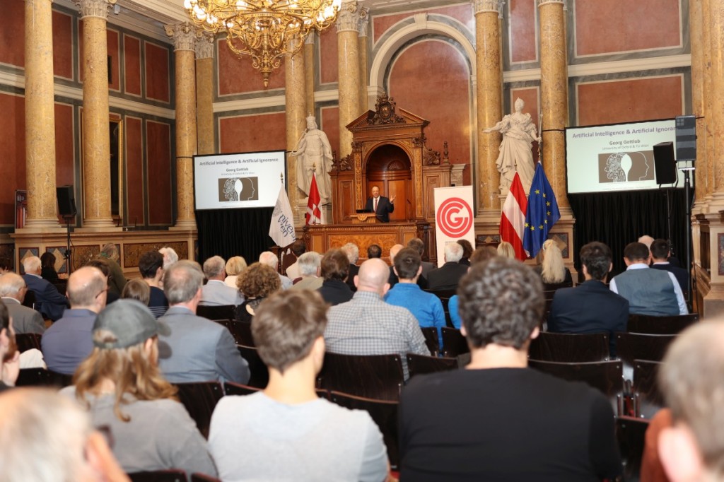Publikum von hinten fotografiert mit Blick auf das Podium im Uni Wien Festsaal, während Georg Gottlob spricht.